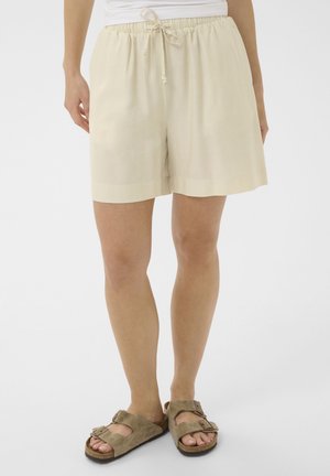 Nő bézs laza rövidnadrágban, húzózsinóros derékrésszel és taupe színű, kétpántos csatos szandálokkal, fehér háttér előtt állva.