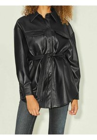 Ylimitoitettu musta keinonahkainen paitajakku, jossa on vyötärölle kevyt vyö, kaksi etutaskua ja napillinen etuosa. Sileä pinta ja kiiltävä viimeistely.