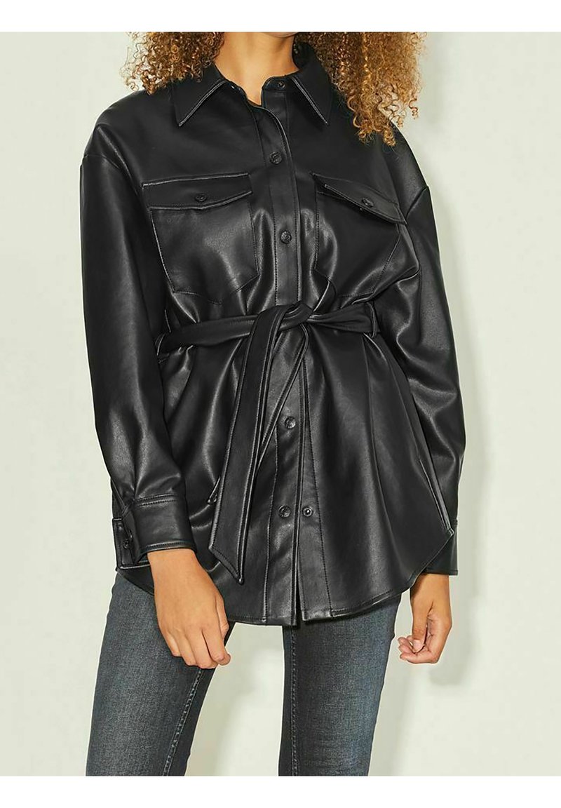 Ylimitoitettu musta keinonahkainen paitajakku, jossa on vyötärölle kevyt vyö, kaksi etutaskua ja napillinen etuosa. Sileä pinta ja kiiltävä viimeistely.