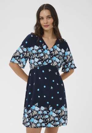 Žena oblečená v námorníckom modrom šate s svetlomodrými a bielymi kvetinovými vzormi, krátkymi rukávmi a výstrihom do V, stojaca s rukami za chrbtom.