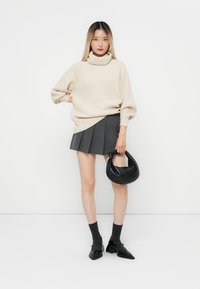 Cremfarbener gerippter Rollkragenpullover, grauer plissierter Rock, schwarze Handtasche, graue Socken und schwarze Schuhe. Das Model steht vor einem weißen Hintergrund.