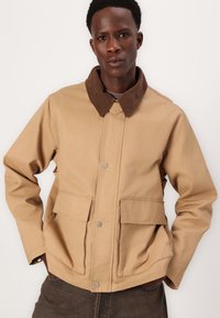 Tan cotton jacket with a brown corduroy collar and snap-button closure. Features two front pockets and a relaxed fit design.