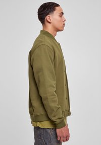 Ung mand i profil iført en olivenfarvet bomberjakke over en lysere grøn skjorte og blå denimjeans foran en ensfarvet grå baggrund.