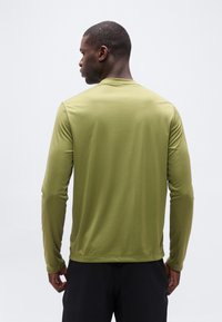 Άνδρας που στέκεται με την πλάτη προς τα εμάς, φοράει μακρυμάνικο πουκάμισο χακί και μαύρο παντελόνι, μπροστά σε απλό ανοιχτόχρωμο φόντο.