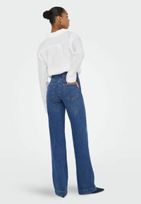 Jeans s vysokým pasem a širokými nohavicemi v modré barvě s klasickým designem a zadními kapsami, v kombinaci s volně střiženou bílou košilí s dlouhým rukávem.