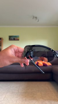 Fekete napszemüveg, ívelt kerettel és színezett lencsékkel. A szár tapintásos fogantyúval rendelkezik a kényelem érdekében. A háttér egy semleges szoba.