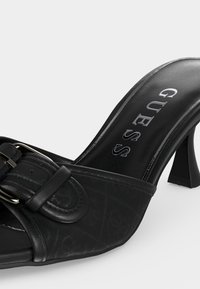 Mule à talon ouverte noire avec motif embossé et boucle décorative sur la bride, avec le nom de la marque « GUESS » sur la semelle intérieure.