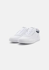 Baskets blanches avec une surface lisse, un design à lacets et des perforations subtiles. Elles présentent un accent de talon marine et un logo sur la languette.