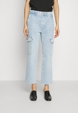 Pantalon cargo en jean bleu clair à jambes droite avec poches à rabat, porté avec un haut noir et des chaussures noires sans lacets.