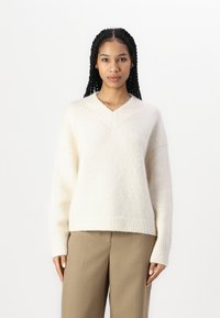 Beige strikket genser med V-hals, avslappet passform, ribbekantede mansjetter og hem, laget av mykt teksturert materiale.