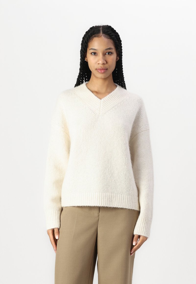 Beige strikket genser med V-hals, avslappet passform, ribbekantede mansjetter og hem, laget av mykt teksturert materiale.