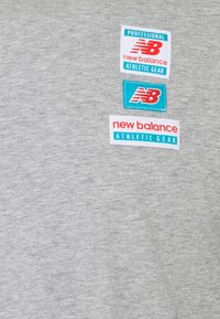 Šedé sportovní tričko s třemi nášivkami s logem "New Balance", bílými a tyrkysovými akcenty a různými styly písma. Hladká textura.