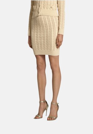 Mujer con falda de punto beige con trenzas y suéter a juego, combinados con sandalias metálicas de tacón alto con tiras en el tobillo y punta abierta, de pie sobre un fondo blanco.