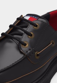 Μαύρο δερμάτινο παπούτσι σκάφους με μαύρα κορδόνια, μπρούτζινα οπές με χαραγμένο το "Tommy" και λογότυπο Tommy Hilfiger σε κόκκινο, λευκό και μπλε στη γλώσσα.