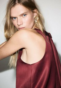 Top burdeos sin mangas con un detalle de espalda drapeada, textura de satén suave y un ligero brillo; combinado con pendientes decorativos de oro.