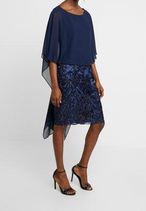 Kobieta ubrana w granatową sukienkę z przezroczystymi rękawami w formie peleryny oraz koronkową spódnicę z perłowymi kwiatowymi zdobieniami, do tego czarne sandały na wysokim obcasie.