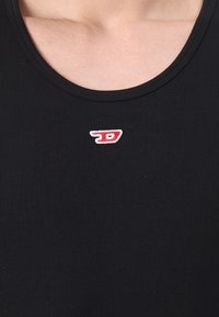Primo piano di un top nero a coste con un piccolo logo ricamato rosso e bianco centrato sotto la scollatura.