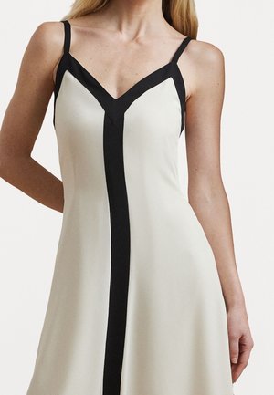 Femme portant une robe blanche sans manches avec des détails de bordure noirs verticaux et diagonaux, debout avec un bras plié derrière le dos.