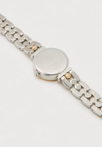 Sat od nehrđajućeg čelika s okruglim srebrnim kućištem, povezanim remenom, zlatnim detaljima i glatkom teksturom. Stražnja strana sadrži ugravirani brend i krunicu.