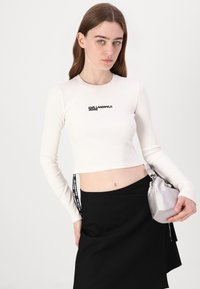 Dugi rukav, rebrasti crop top u boji slonovače s crnim logom "Karl Lagerfeld Jeans"; uparen s crnom suknjom i srebrnom torbom.
