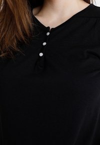 Czarna koszulka w stylu henley, wykonana z miękkiego materiału, z okrągłym dekoltem i trzema perłowymi guzikami z przodu. Gładka faktura z lekkim połyskiem.