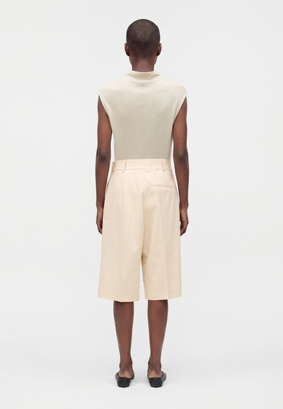 Personne portant un haut beige sans manches, un short crème taille haute longueur genoux, et des chaussures noires à enfiler, debout de dos sur un fond uni.