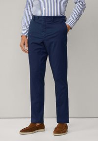 Chinos blu navy con una texture liscia, dotati di passanti per cintura e tasche laterali. Abbinati a scarpe brown slip-on. Camicia a righe visibile sopra.