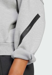 Šedá mikina se strukturovaným, ohnutým manžetem a černým akcentovým pásem na rukávu s textem "SPORTOVNÍ MÓDA." Měkký, ležérní design.