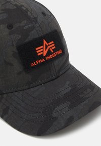Gorra de béisbol con camuflaje gris oscuro, paneles cosidos y una parche negra con el logo rojo de Alpha Industries y texto.