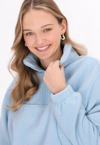 Pullover in pile azzurro chiaro con colletto alto e chiusura a mezza zip. Dettagli notevoli includono una texture morbida e polsini elastici.