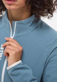 Veste en polaire texturée bleu clair avec un col montant et une fermeture éclair blanche. Présente un logo sur le devant et des poignets de manches contrastants.