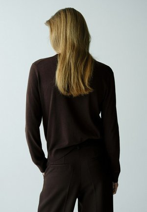 Personne aux longs cheveux blonds portant un haut à manches longues marron foncé et un pantalon assorti, debout les mains dans les poches, de dos.