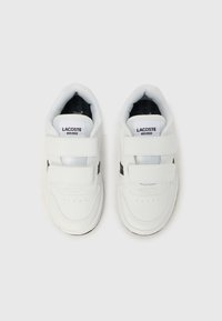 Λευκά sneakers Lacoste με ιμάντες βέλκρο, που διαθέτουν υφασμένο πάτο, στρογγυλεμένη μύτη και μαύρες λεπτομέρειες στα πλάγια. Λογότυπο της μάρκας στη γλώσσα.