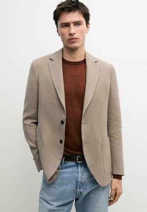 Giovane uomo che indossa un blazer beige sopra un maglione marrone abbinato a jeans azzurri, in piedi davanti a uno sfondo bianco uniforme.