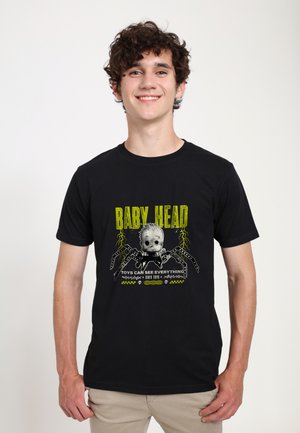 Giovane uomo sorridente che indossa una maglietta nera con una grafica di ragno e testo giallo con la scritta "Baby Head, Toys Can See Everything."