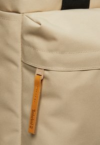 Mochila de tela beige con un acabado texturizado, que cuenta con un bolsillo lateral y una pestaña de cuero con el logotipo "Timberland" grabado.