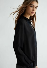 Blusa nera a maniche lunghe con colletto resistente, caratterizzata da una texture liscia e scollatura a V, per una silhouette slanciata.