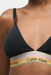 Κοντινό πλάνο ενός ατόμου που φοράει μαύρο bralette Calvin Klein με λουρί ουράνιου τόξου σε διαβαθμισμένα χρώματα και λεπτούς ιμάντες ώμου.