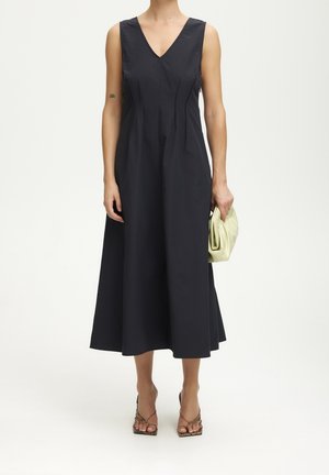 Robe midi noire sans manches à encolure en V portée avec des talons ouverts, tenant un sac à main plié vert clair sur un fond blanc uni.