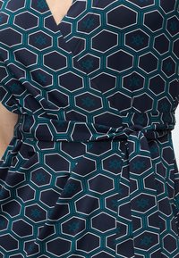 Ναυτικό φόρεμα τύπου wrap με σχέδιο από τιρκουάζ και λευκά εξάγωνα και γεωμετρικά αστέρια, διαθέτει δεμένη ζώνη στη μέση.