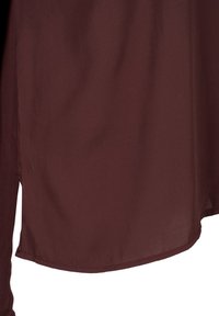 Tissu bordeaux foncé avec un ourlet incurvé et une petite fente latérale, montrant la texture et les détails des coutures.