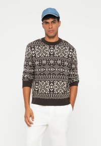 Brauner Strickpullover mit weißen geometrischen Mustern, rundem Halsausschnitt und gerippten Bündchen. Kombiniert mit hellen Hosen und einer Jeanskappe.
