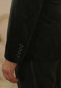 Schwarze Wildleder-Blazerärmel mit vier schwarzen Knöpfen, glatter Textur und körperbetonter Passform. Die Hand ruht sanft an der Seite.