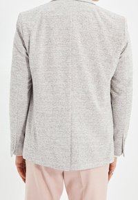 Blazer grigio a fantasia, tessuto testurizzato, con uno spacco singolo sul retro, maniche lunghe con dettagli a bottoni, abbinato a pantaloni rosa chiaro.