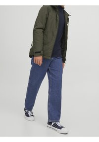 Persona in cammino con indosso una giacca verde oliva, una camicia blu navy, jeans blu e sneakers bianco e nere su uno sfondo neutro.