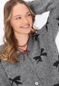 Cardigan grigio lavorato a maglia con motivi a fiocco neri, dotato di chiusura a bottoni. Collana a strati in rosso con un ciondolo a forma di cuore. Tessuto morbido e testurizzato.