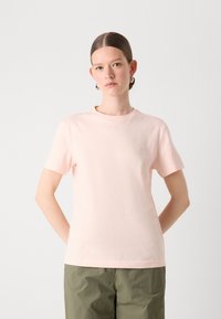 T-shirt in cotone rosa chiaro con maniche corte, scollatura a girocollo e dettaglio logo discreto sul petto. Indossato con pantaloni verde oliva.