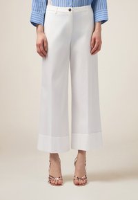 Pantalons blancs à jambes larges avec une texture lisse, présentant un ourlet retroussé et une fermeture à un bouton. Associés à des sandales à lanières métalliques.