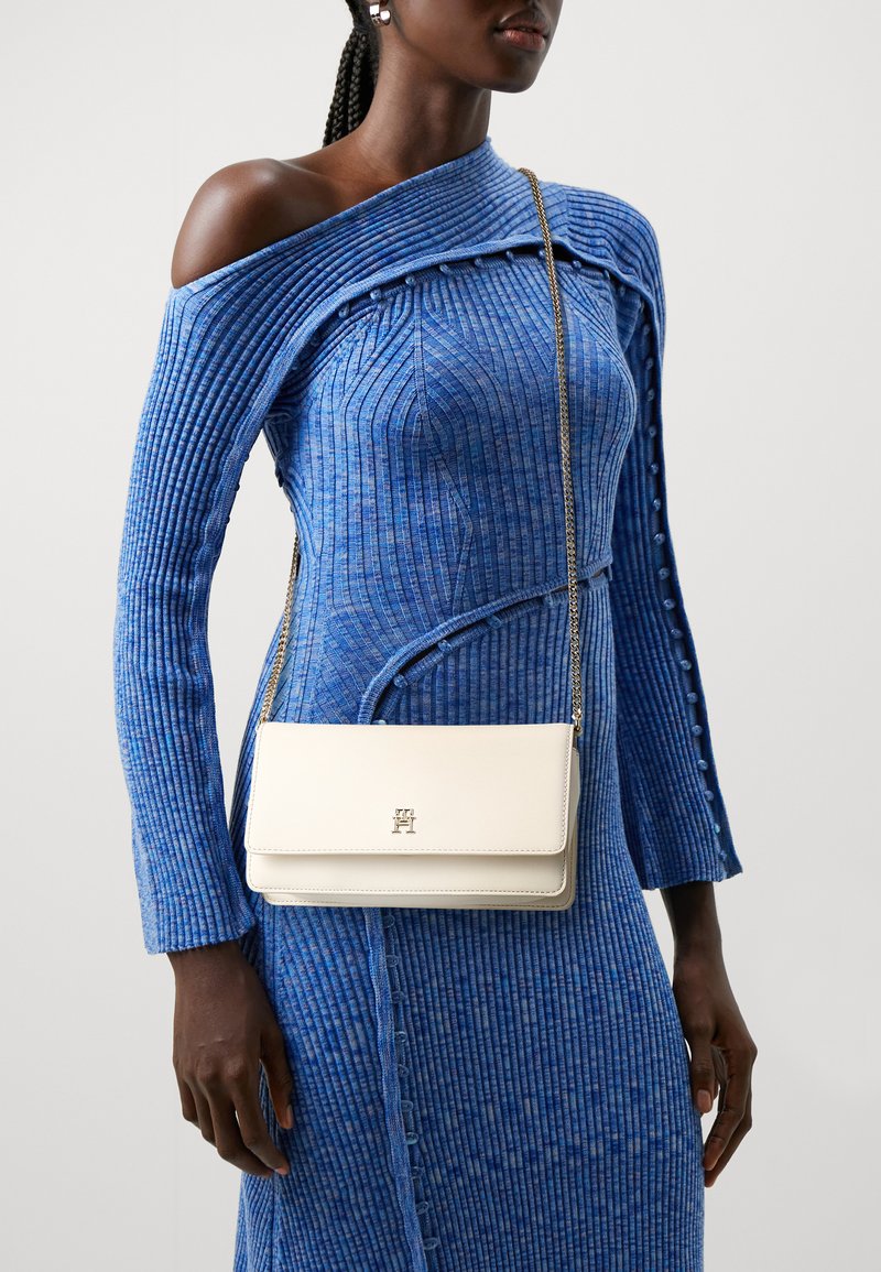Rochie tricotată albastră, cu design off-shoulder; geantă crossbody din piele albă, cu un logo auriu și curea metalică.