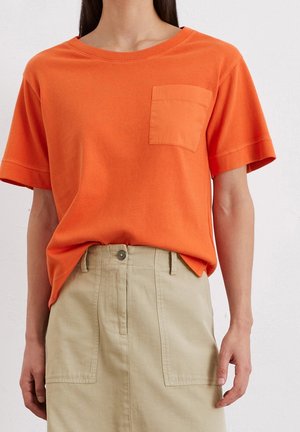 Osoba ubrana w jasnopomarańczową koszulę z krótkim rękawem i kieszenią na piersi oraz beżową spódnicę z wysokim stanem i przednimi kieszeniami.